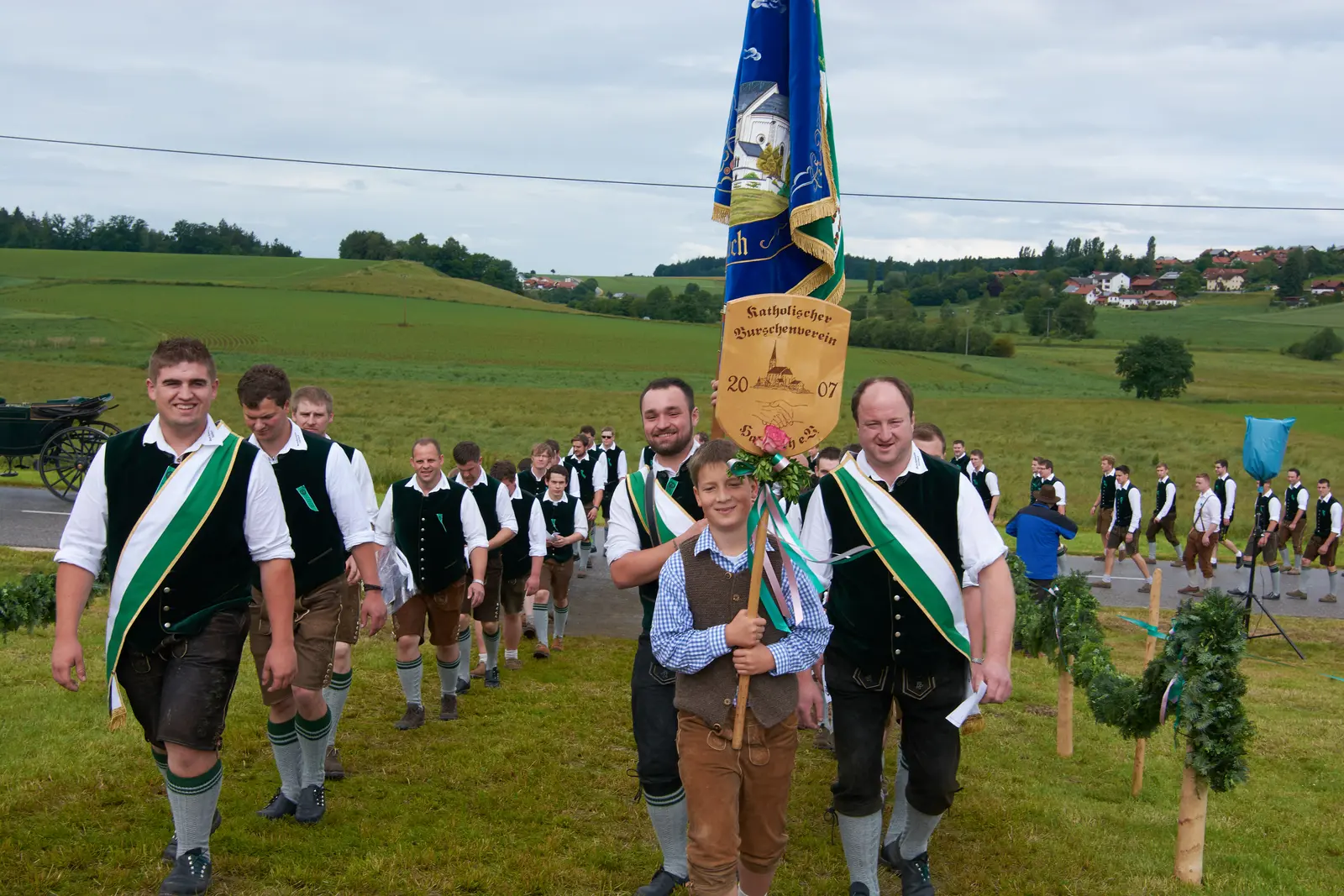 Der Verein zieht in Tracht mit grüner Schärpe über eine Wiese, vorneweg ein Bub mit der Vereinstafel, dahinter die Fahne.
