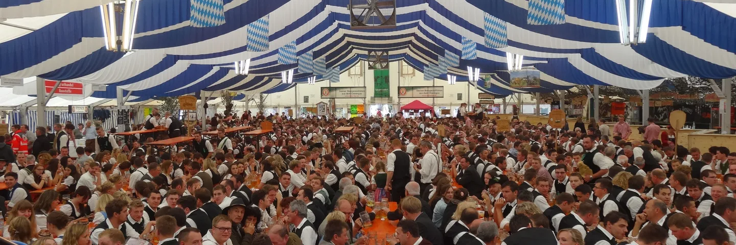 Voll besetztes Festzelt mit blau-weißem Himmel, an den Bierbänken sitzen Burschen und Dirndl in Tracht