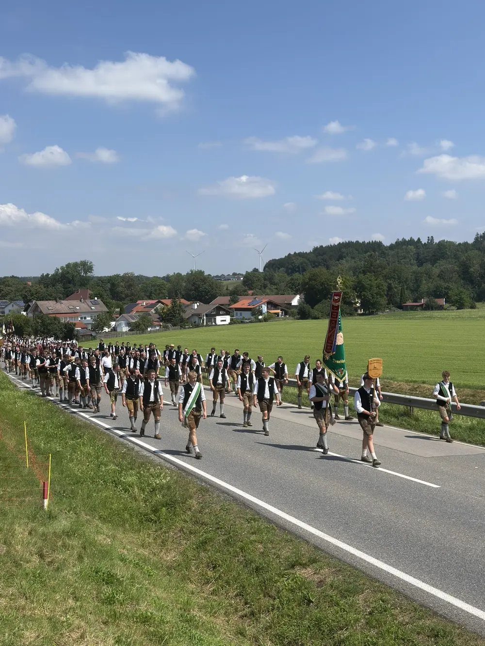 Langer Festzug in Tracht auf der Landstraße, Felder und Ort im Hintergrund