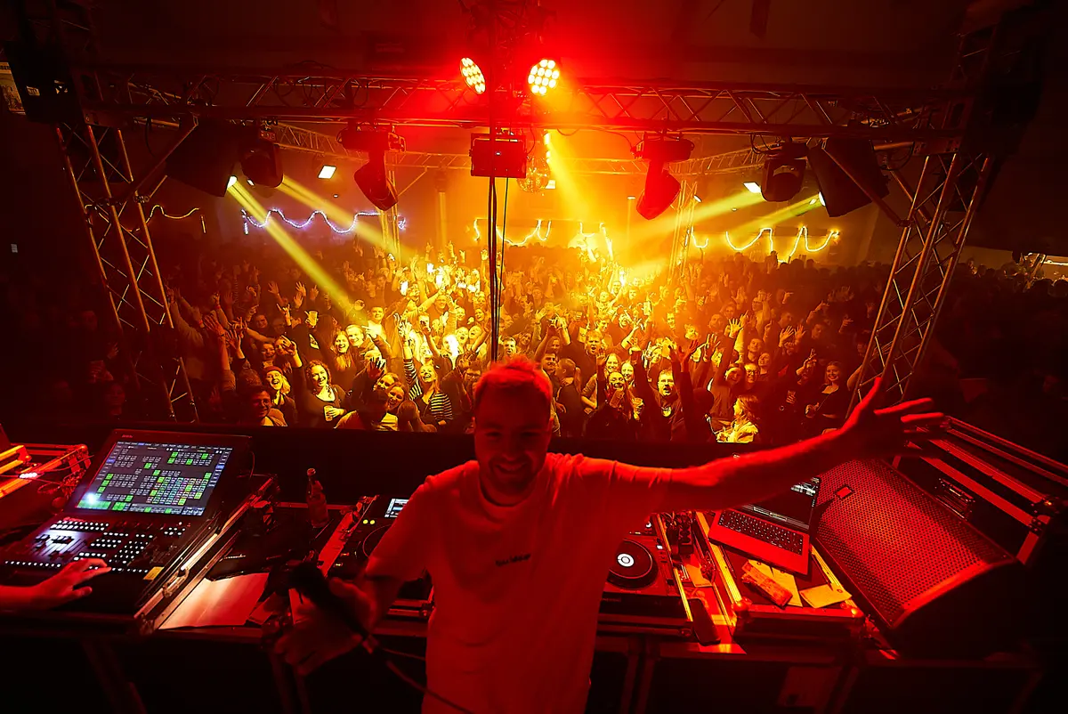 Der DJ steht mit ausgebreiteten Armen hinter dem Pult, davor die feiernde Menge