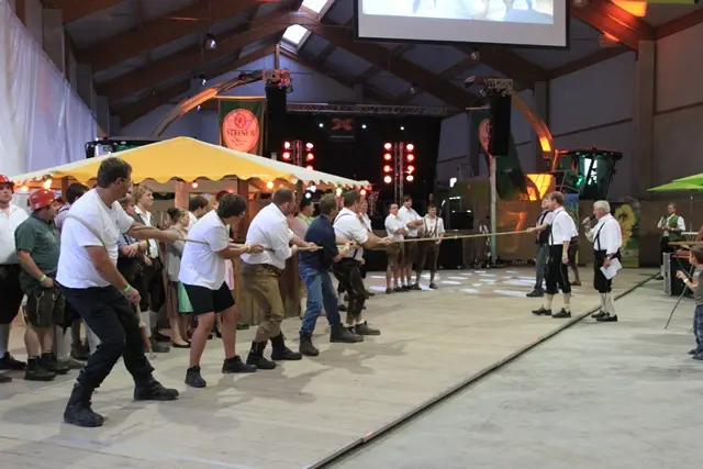 Eine Mannschaft in Lederhosen zieht am Seil, dahinter Festzelt, Bühne und Publikum in der Maschinenhalle.