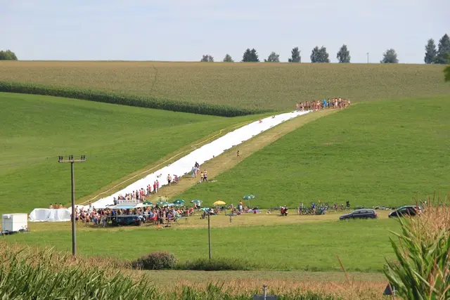 Die Wasserrutschn zieht sich als weiße Bahn den Wiesenhang hinunter, Zuschauer am Rand