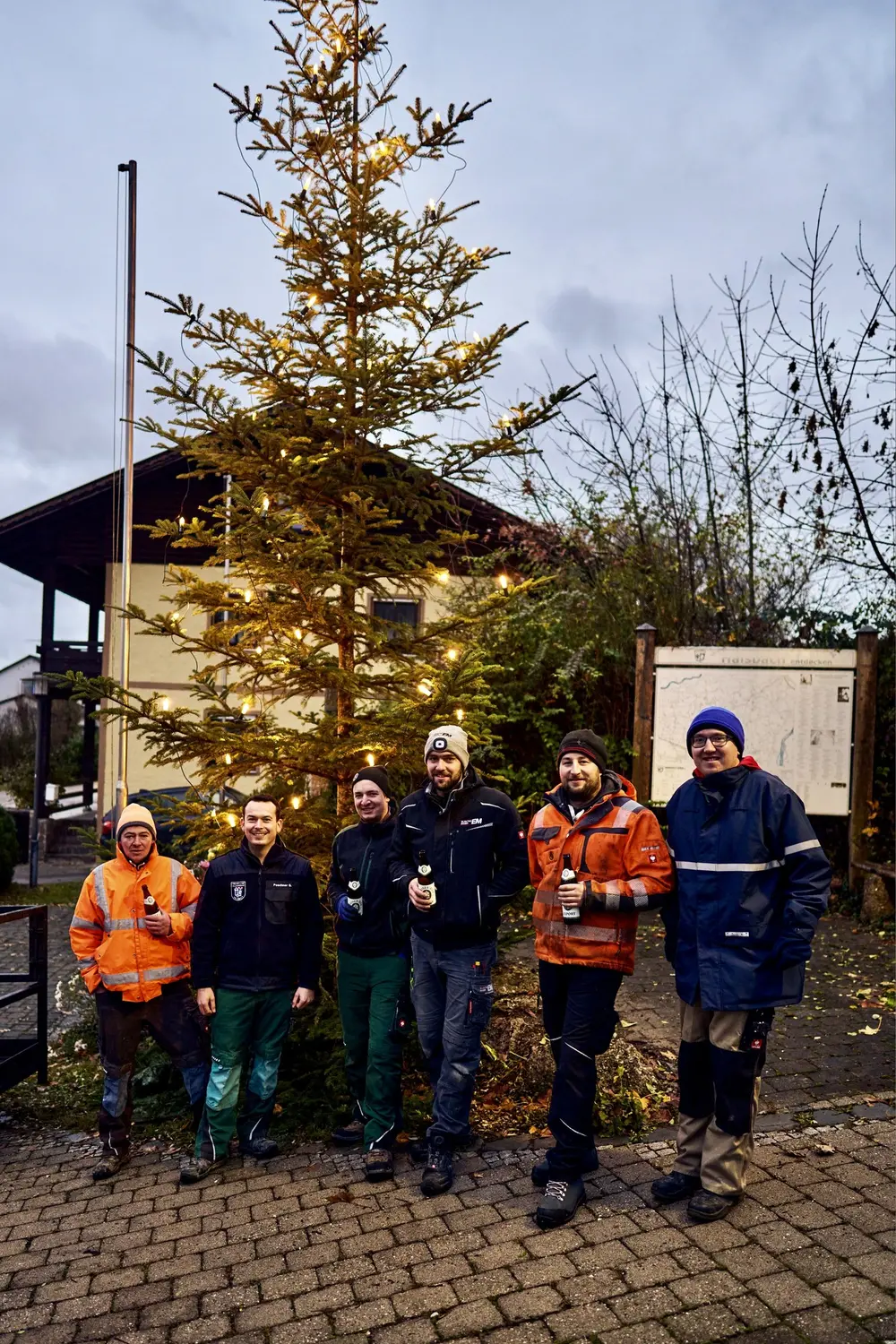 Sechs Burschen in Arbeitskleidung mit Bechern in der Hand vor dem frisch aufgestellten Christbaum am Dorfplatz